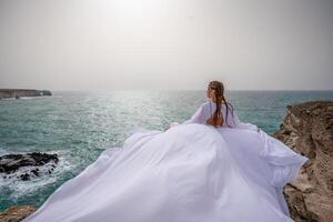 contento la libertà donna su il spiaggia godendo e in posa nel bianca vestire. posteriore Visualizza di un' ragazza nel un' svolazzanti bianca vestito nel il vento. vacanze, vacanze a mare. foto