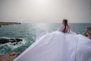 contento la libertà donna su il spiaggia godendo e in posa nel bianca vestire. posteriore Visualizza di un' ragazza nel un' svolazzanti bianca vestito nel il vento. vacanze, vacanze a mare. foto