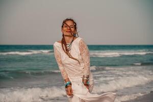 modello nel boho stile nel un' bianca lungo vestito e argento gioielleria su il spiaggia. sua capelli è intrecciato, e Là siamo molti bracciali su sua braccia. foto