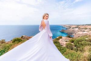 bionda con lungo capelli su un' soleggiato riva del mare nel un' bianca fluente vestire, posteriore Visualizza, seta tessuto agitando nel il vento. contro il fondale di il blu cielo e montagne su il spiaggia. foto