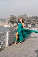 donna mare verde vestire. lato Visualizza un' contento donna con lungo capelli nel un' lungo menta vestito in posa su un' spiaggia con calma mare bokeh luci su soleggiato giorno. ragazza su il natura su blu cielo sfondo. foto