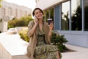 all'aperto foto di elegante bellissimo donna con corto taglio di capelli godendo autunno,n giorno Tenere tazza di caffè, utilizzando mobile Telefono. usurante casuale jacker e elegante vestire.