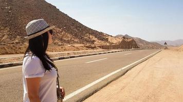 una ragazza bianca con un cappello si trova vicino a un'ampia strada asfaltata deserta. ritratto posteriore di una donna che guarda in lontananza su una strada deserta con montagne e sole caldo. vicino Oriente. concetto di viaggio foto