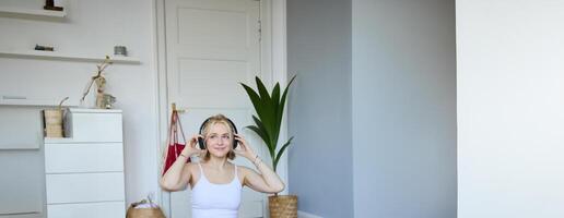 verticale tiro di sorridente giovane donna utilizzando tutorial per allenarsi a casa, seduta con il computer portatile nel senza fili cuffie su yoga stuoia, segue fitness istruttore su sociale media, ripete esercizi foto