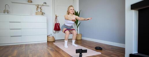 ritratto di donna, fitness istruttore a casa, registrazione di allenamento, mostrando Come per fare gamba esercizi, squat con elastico resistenza gruppo musicale, Lavorando su in casa su yoga stuoia foto