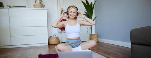 ritratto di in forma e salutare giovane donna nel senza fili cuffia, fa allenamento, utilizzando in linea tutorial, ascolta per fitness istruttore su computer portatile, esercizio a casa foto