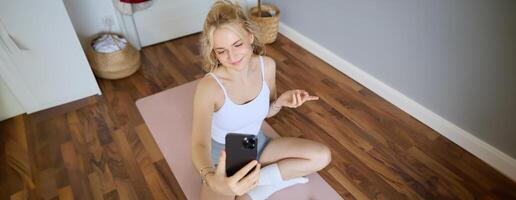 ritratto di donna avendo vivere ruscello formazione sessione, utilizzando sua smartphone per creare allenarsi contenuto, blogging durante esercizi o fare yoga, seduta su gomma da cancellare stuoia a casa foto