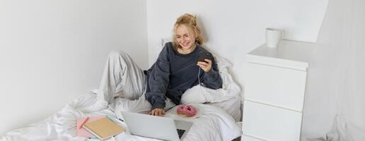 stile di vita e persone concetto. giovane bellissimo donna, soggiorno a casa, dire bugie nel letto con il computer portatile e smartphone, mangiare ciambella, godendo gratuito volta, la spesa fine settimana a casa, Guardando film in linea foto