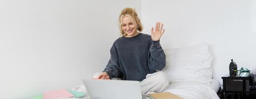 ritratto di giovane contento donna, si connette per Chiacchierare, utilizzando computer portatile, agitando mano a telecamera, detto Ciao per qualcuno in linea, seduta su letto, studiando, e-learning o Lavorando a partire dal casa foto