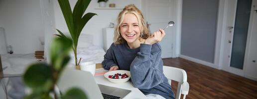 Immagine di ridendo donna seduta nel davanti di il computer portatile nel sua camera, mangiare colazione, Tenere cucchiaio e ciotola nel mano, Guardando S in linea mentre avendo un' merenda foto