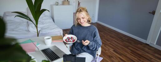 ritratto di sorridente giovane femmina vlogger, registrazione e la creazione di soddisfare nel sua camera, mangiare nel davanti di telecamera, parlando, fabbricazione stile di vita blog foto