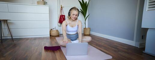 ritratto di donna durante allenamento, seduta su yoga stuoia con resistenza gruppo musicale, ascoltando per Istruzioni su computer portatile, indossare senza fili cuffia, ripetendo esercizi foto