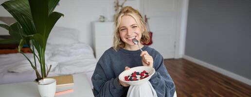 ritratto di carino sorridente biondo donna mangia prima colazione nel sua Camera da letto, guardare a telecamera, Tenere ciotola con dolce e cucchiaio foto