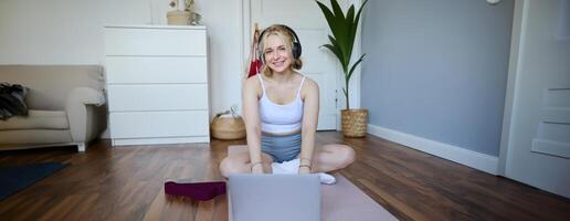 ritratto di donna durante allenamento, seduta su yoga stuoia con resistenza gruppo musicale, ascoltando per Istruzioni su computer portatile, indossare senza fili cuffia, ripetendo esercizi foto