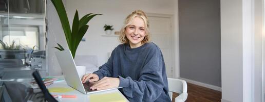 ritratto di sorridente giovane donna, Università alunno si siede nel sua camera, fa compiti a casa, studi remoto a partire dal casa, usi il computer portatile per libero professionista opera foto