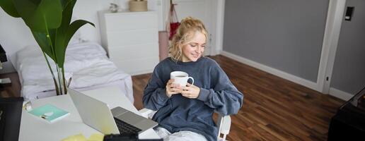 ritratto di giovane sorridente donna, vlogger registrazione stile di vita nel sua camera, si siede nel davanti di il computer portatile e digitale telecamera, bevande Tè, crea soddisfare per sociale media account foto