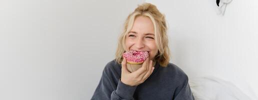 vicino su ritratto di felice, carino biondo donna, Tenere ciambella, mangiare dolce, delizioso comfort cibo, mostrando dolce a telecamera foto