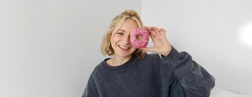 vicino su ritratto di felice, carino biondo donna, Tenere ciambella, mangiare dolce, delizioso comfort cibo, mostrando dolce a telecamera foto