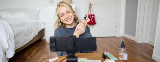 ritratto di giovane creativo sociale media soddisfare Creatore, donna mostrando rossetto campioni su sua mano, registrazione di bellezza e trucco, seduta nel sua camera nel davanti di digitale telecamera foto