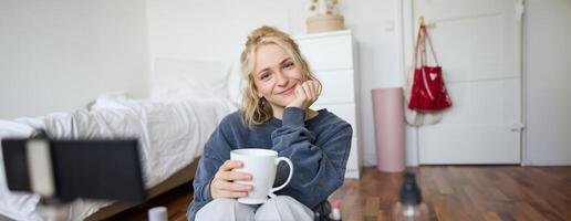 vicino su ritratto di biondo giovane donna, seduta nel sua camera, guardare a telecamera, Tenere tazza di Tè, registrazione accogliente stile di vita blog, vlogger la creazione di soddisfare per sociale media account foto