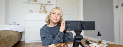 ritratto di giovane creativo sociale media soddisfare Creatore, donna mostrando rossetto campioni su sua mano, registrazione di bellezza e trucco, seduta nel sua camera nel davanti di digitale telecamera foto