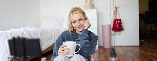 vicino su ritratto di bellissimo biondo donna, ragazza si siede nel sua camera, fa trucco, record per sociale media, bevande Tè, detiene boccale e sorrisi a telecamera foto