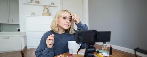 ritratto di Bellissima, sorridente biondo donna, ragazza registrazione di sua trucco lezione per sociale media, vlogger seduta su pavimento nel sua camera, utilizzando stabilizzatore per creare contenuto, rivedere mascara foto