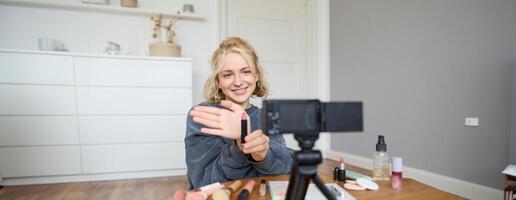 Immagine di giovane femmina soddisfare Creatore, bellezza blogger si siede su pavimento nel sua camera, record di trucco, Spettacoli rossetto su telecamera foto