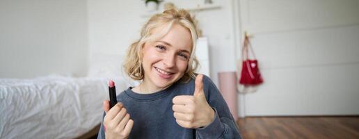 carino sorridente ragazza Spettacoli rossetto e pollici su mano cartello, consigliare bellezza Prodotto, record soddisfare per sociale media account, stile di vita video blog foto