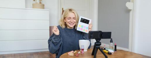 eccitato sorridente biondo donna registrazione revisione su un' nuovo ombretto tavolozza, stile di vita blogger mostrando Prodotto per seguaci, si siede su pavimento con entusiasta viso foto