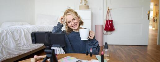 ritratto di biondo giovane stile di vita blogger, donna record di sua parlando di vita e bellezza, si siede nel davanti di telecamera, detiene tazza, bevande Tè, fa blog soddisfare per sociale media account foto