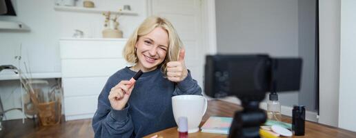 ritratto di sorridente, bellissimo giovane donna, stile di vita blogger registrazione un' nel sua camera, seduta nel davanti di telecamera su stabilizzatore, mostrando pollici su e occhiolino viso, consigliare mascara foto