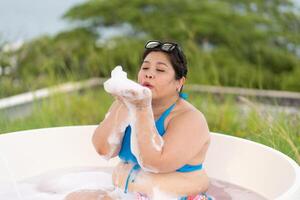 asiatico più dimensione donna indossare costumi da bagno giocando sapone bolla nel bagno vasca durante vacanza foto