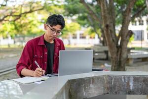 asiatico Università alunno studiando su il computer portatile a città universitaria all'aperto parco. uomo scrittura su un' Nota libro e Lavorando su il computer portatile. educativo concetto foto