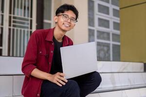 ritratto di asiatico Università alunno utilizzando il computer portatile guardare per telecamera. un' uomo Lavorando con un' il computer portatile computer a città universitaria foto