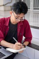 asiatico Università alunno studiando su il computer portatile a città universitaria all'aperto parco. uomo scrittura su un' Nota libro e Lavorando su il computer portatile. educativo concetto foto