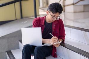 asiatico Università alunno studiando su il computer portatile a città universitaria all'aperto parco. uomo scrittura su un' Nota libro e Lavorando su il computer portatile. educativo concetto foto