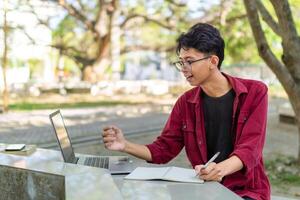 asiatico Università alunno studiando su il computer portatile a città universitaria all'aperto parco. uomo scrittura su un' Nota libro e Lavorando su il computer portatile. educativo concetto foto