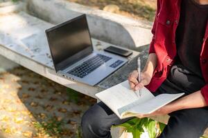 asiatico Università alunno studiando su il computer portatile a città universitaria all'aperto parco. uomo scrittura su un' Nota libro e Lavorando su il computer portatile. educativo concetto foto