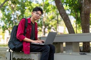 ritratto di asiatico Università alunno utilizzando il computer portatile guardare per telecamera. un' uomo Lavorando con un' il computer portatile computer a città universitaria foto