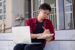 asiatico Università alunno studiando su il computer portatile a città universitaria all'aperto parco. uomo scrittura su un' Nota libro e Lavorando su il computer portatile. educativo concetto foto