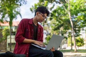 ritratto di asiatico Università alunno utilizzando il computer portatile guardare per telecamera. un' uomo Lavorando con un' il computer portatile computer a città universitaria foto