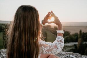 contento donna nel bianca boho vestito fabbricazione cuore cartello con mani su tramonto nel montagne. romantico donna con lungo capelli in piedi con sua indietro su il tramonto nel natura nel estate con Aperto mani. foto