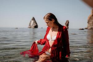 donna viaggio mare. contento turista nel rosso vestito godere assunzione immagine all'aperto per ricordi. donna viaggiatore in posa nel mare spiaggia, circondato di vulcanico montagne, condivisione viaggio avventura viaggio foto