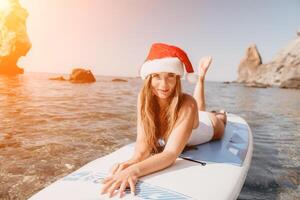 donna mare sup. vicino su ritratto di contento giovane caucasico donna con lungo capelli nel Santa cappello guardare a telecamera e sorridente. carino donna ritratto nel un' bianca bikini in posa su cenare tavola nel il mare foto