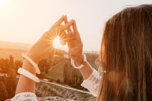 contento donna nel bianca boho vestito fabbricazione cuore cartello con mani su tramonto nel montagne. romantico donna con lungo capelli in piedi con sua indietro su il tramonto nel natura nel estate con Aperto mani. foto