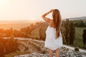 contento donna in piedi con sua indietro su il tramonto nel natura nel estate con Aperto mani. romantico bellissimo sposa nel bianca boho vestito in posa con montagne su tramonto foto