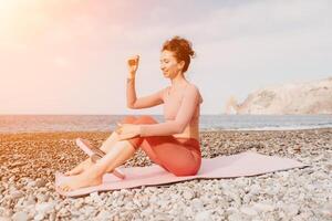 donna mare pilates. sportivo contento mezzo anziano donna praticante fitness su spiaggia vicino mare, sorridente attivo femmina formazione con squillare su yoga stuoia fuori, godendo salutare stile di vita, armonia e meditazione foto