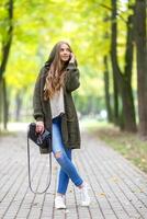 bellissimo giovane donna nel verde a maglia cardigan con un' cappuccio a piedi nel autunno parco e parlando mobile Telefono. autunno alla moda concetto. foto