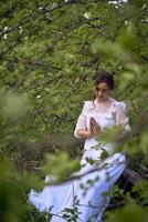 un' bellissimo donna nel un' bianca Vintage ▾ vestito con un' treno è preghiere mentre seduta su un' caduto albero foto
